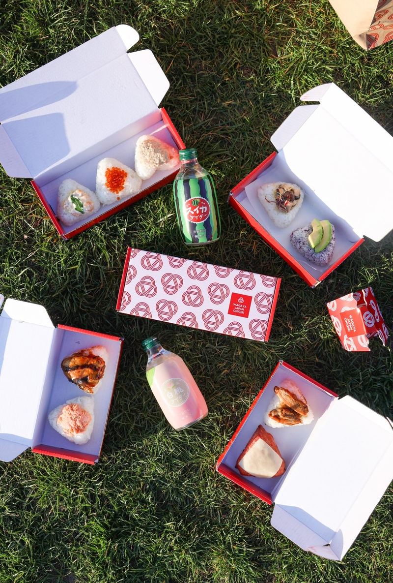 Wagaya red lunch boxes with a selection of onigiri arranged on grass for an outdoor gathering